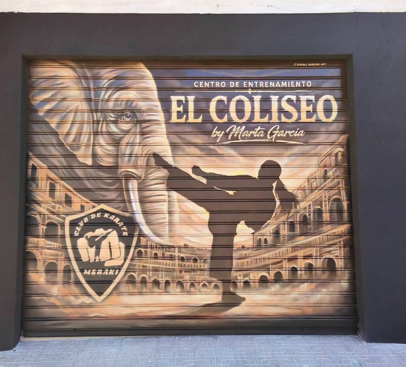 Fachada real de El Coliseo en Almendralejo: puerta pintada con el elefante del emblema, una karateca y el anfiteatro, junto al escudo del Club de Karate Meraki.